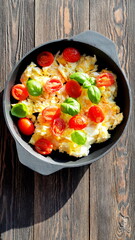 Scrambled eggs with slices of fresh cherry tomatoes and basil leaves in a cast-iron pan on a wooden table under the sun's rays.