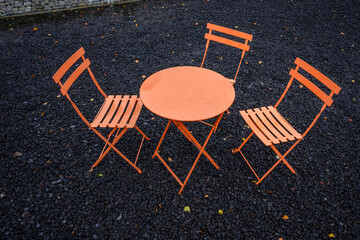 Orange bistro table and three chairs outside on a wet day, black gravel 

