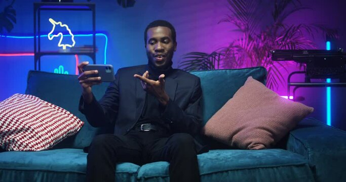 Joyful handsome stylish African American man in black clothes sitting on couch and havong conversation via smartphone. Young guy videochatting on phone in room with neon lights. Male talking on