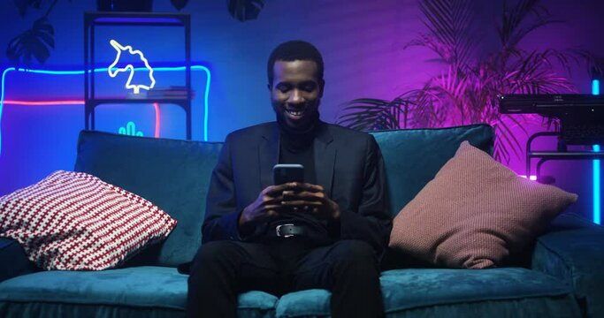 Smiled cheerful good looking stylish African American man in black clothes sitting on couch. Handsome guy using smartphone on sofa in living room with neon lights. Male tapping on phone while chatting