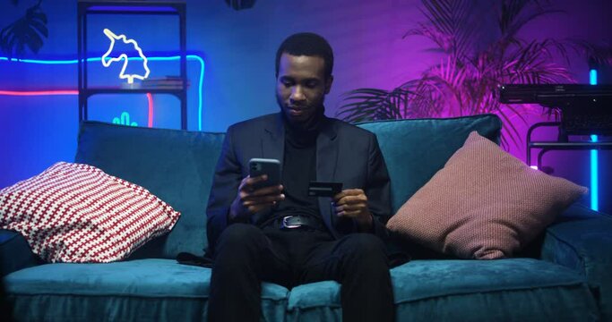 Handsome young African American stylish man sitting on couch and shopping online on smartphone in living room with neon light. Black guy tapping on his phone and holding credit card as paying for buy
