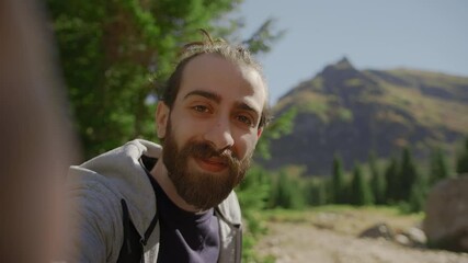 White toothed smile face hippie climber with his backpack behind him, with his handheld camera or phone, salutes the audience or the person he is talking to and points to a high mountain. 