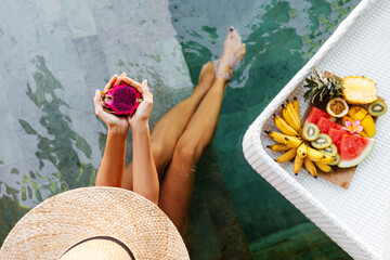 Woman holding dragon fruit near tray with served Breakfast in swimming pool. Tropical exotic fruits on wooden plate