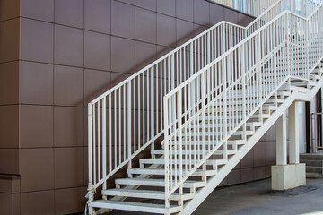 White wide metal pedestrian staircase with handrails at a city building with a brown tiled wall. Steel framed staircase for access to upper floor. Daylight urban background with place for copy space.