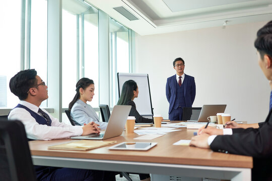 Asian Manager Speaking To Group Of Business People During Team Meeting In Office