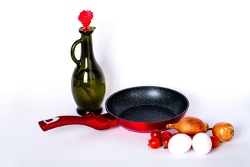 Two eggs, glass carafe of vegetable oil, onions, red frying pan and cherry tomatoes on a white...