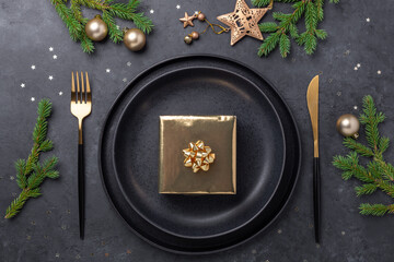 Obraz na płótnie Canvas Christmas table setting. Black ceramic plate with golden gift box, fir tree branch and accessories on stone background. Gold decoration