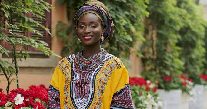 Portrait shot of the beautiful and happy Afrcan woman in the traditional outfit standing in the cozy courtyard with flowers and smiling to the camera. Outdoor.
