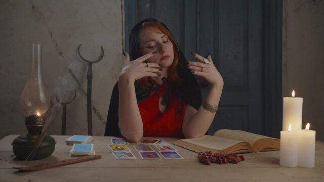 Mysterious red haired soothsayer in black cape sitting at rustic table with book of magic spell and tarot cards, fortune telling and interpreting future with runic stones in dimmed rural house.