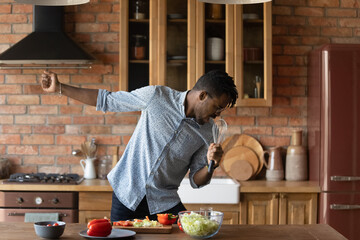 Happy young African American man have fun preparing healthy delicious breakfast in modern home kitchen. Overjoyed biracial male tenant sing in appliance enjoy good morning cooking food in own house. - Powered by Adobe