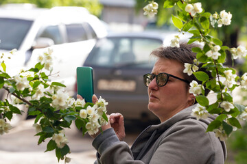 A short-haired woman in a gray sweatshirt takes pictures of white flowers on a bush with her phone. A warm spring day, a walk around the city.