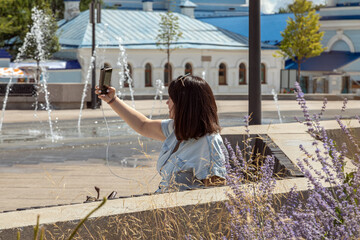 A young woman with a phone sits on a bench near a fountain in a city park. Clear, warm day, selective focus.