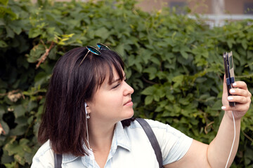 A young woman takes a selfie against the background of a green bush in a city park. Summer warm day, city walks, close-up.
