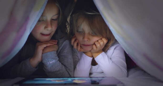 Child blonde caucasian girl with friend or sister watching movie or animation on tablet app under bed blanket at night. Modern technology childhood kid use at home. 4k video