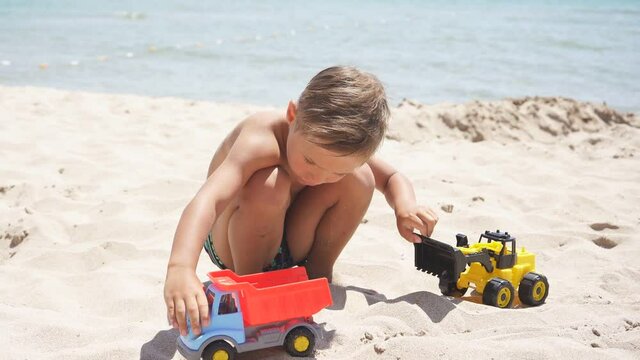 Cute child, boy playing with cars on a sandy beach, plastic toy car, excavator. A child and the azure sea on a summer day.