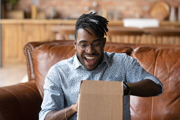 Overjoyed young African American man unpack cardboard box triumph with good quality order. Happy...