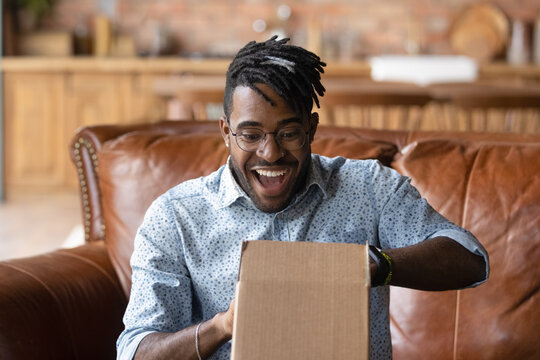 Overjoyed Young African American Man Unpack Cardboard Box Triumph With Good Quality Order. Happy 20s Biracial Male Unbox Carton Package With Internet Delivery, Shopping On Web Online From Home.