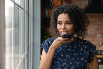 Fototapeta na wymiar Young African American woman stand near window at home record audio message on smartphone. Millennial female talk on loudspeaker or activate digital voice assistant app on modern cellphone gadget.