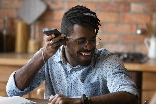 Happy young Africana American man in glasses have fun listen to audio message on smartphone chatting online. Smiling millennial 20s male talk using voice virtual digital assistant app on cellphone. - Powered by Adobe