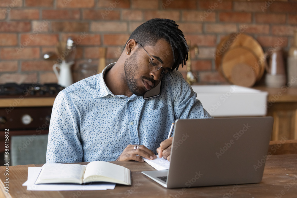 Sticker busy young african american man sit at home kitchen office work distant on laptop talk on cellphone.