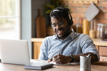 Smiling young African American man in headphones glasses make note watching webinar on laptop....