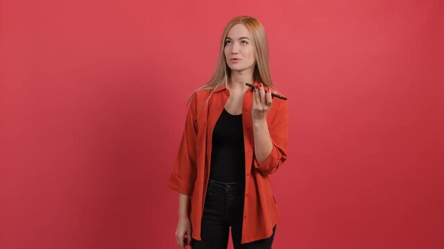 Portrait of a young woman holding her smartphone near her mouth while recording a vocal massage and looking aside, against a red background. The girl talks through the application with his friends.