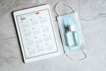 a tablet with an open calendar for 2021 year and protective medical mask and hand sanitizer on a gray concrete background. covid-19 coronavirus protection concept in 2021