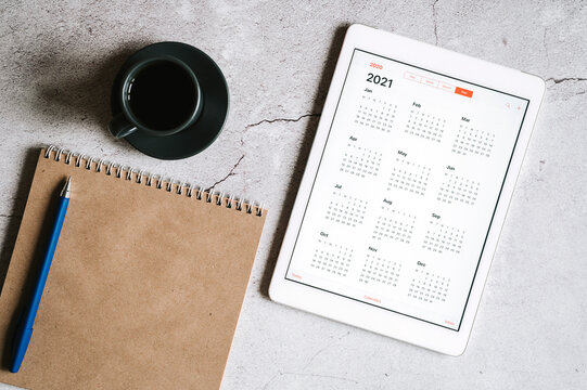 a tablet with an open calendar for 2021 year, a cup of coffee and a craft paper notebook on a gray concrete background