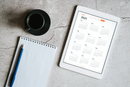 a tablet with an open calendar for 2021 year, a cup of coffee, and a spring notebook with a pen on a gray concrete background