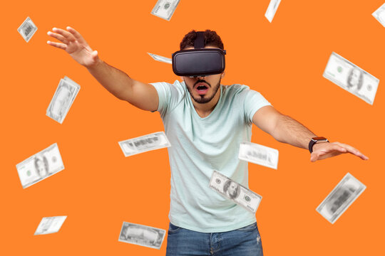 Portrait of shocked man wearing vr glasses, stretching hands forward, trying to get money, dollar rain falling, illusion of rich millionaire, playing virtual reality game, indoor studio shot