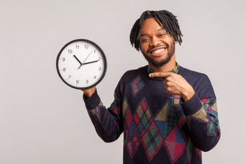Happy satisfied african guy with dreadlocks in casual sweatshirt pointing finger at wall clock with...