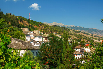 Fototapeta na wymiar Gjirokaster town in Albania – traditional white houses with stone gray roofs on the hill. Summer landscape with lush foliage, blue sky and mountains on the horizon. 