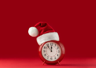 Red alarm clock in Santa Claus hat on red background. Five minutes to midnight. Minimal concept of...