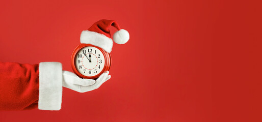 Santa Claus hand holding red alarm clock in Santa hat. Red background. Concept of coming Christmas...