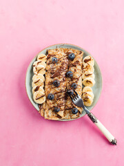 Delicious crepes filled with chocolate hazelnut spread and topped with banana and blueberries, top view on pink background.