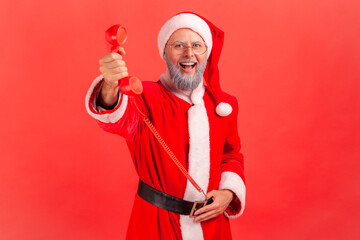 Fototapeta na wymiar Positive smiling man in santa claus costume holding handset of landline phone, offering to call to relatives and congratulate with holidays. Indoor studio shot isolated on red background