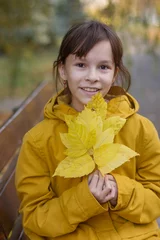 Fotobehang Leuk meisje in gele jas zittend op de bank. Het meisje houdt een armvol bladeren vast © Albert Ziganshin