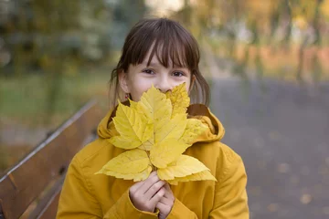 Poster Leuk meisje in gele jas zittend op de bank. Het meisje houdt een armvol bladeren vast © Albert Ziganshin
