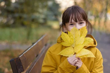 Foto op Canvas Leuk meisje in gele jas zittend op de bank. Het meisje houdt een armvol bladeren vast © Albert Ziganshin