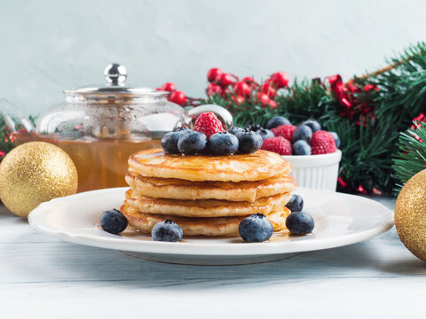 Stack of pancakes with blueberries and maple syrup served in a dish with green tea with Christmas decor on white wooden table
