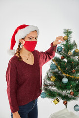 Young brunette Spanish woman dressed in Santa hat celebrating Christmas with gifts and decorating the Christmas tree, covid-19, with a red facing mask