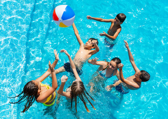 Group of many kids jump after ball and play in swimming pool, splashing, lift hands smile view from...