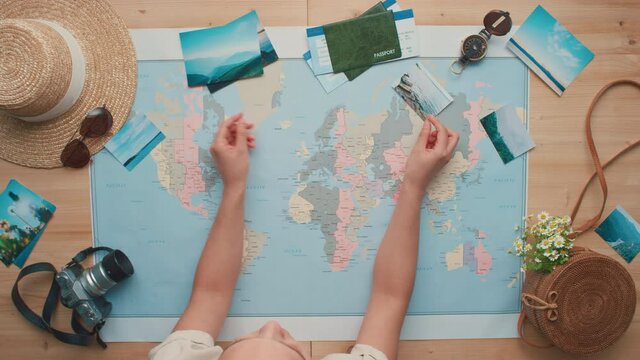 Horizontal top-view flat lay footage of unrecognizable woman holding printed photos of beautiful landmarks in hands laying them on world map