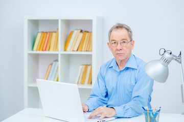 A senior citizen in glasses looks intently at the camera while sitting at the computer at home. Elderly man works remotely from home during the coronavirus pandemic