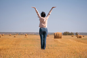 A happy, free, independent, dreamy girl in the field raised her hands to the sky. A woman in a jacket and jeans at the harvest rear view. Silhouette of a girl from behind.Harvesting of grain crops