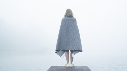 Lonely slender young girl wrapped in a warm gray blanket stands on a wooden cliff near the lake and...
