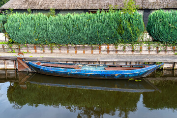 An old blue wooden boat, moored to the shore, with a hedge and thatched roof in the background.