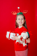 Portrait of a funny cheerful girl with a bandage of horns on her head hugging a teddy bear in Christmas pajamas isolated on a bright red background.