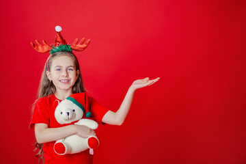 Portrait of a funny cheerful girl with a bandage of horns on her head hugging a teddy bear in Christmas pajamas isolated on a bright red background.