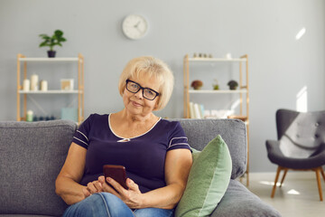 Obraz premium Mature woman sitting on sofa at home, holding mobile phone in hands and looking at camera